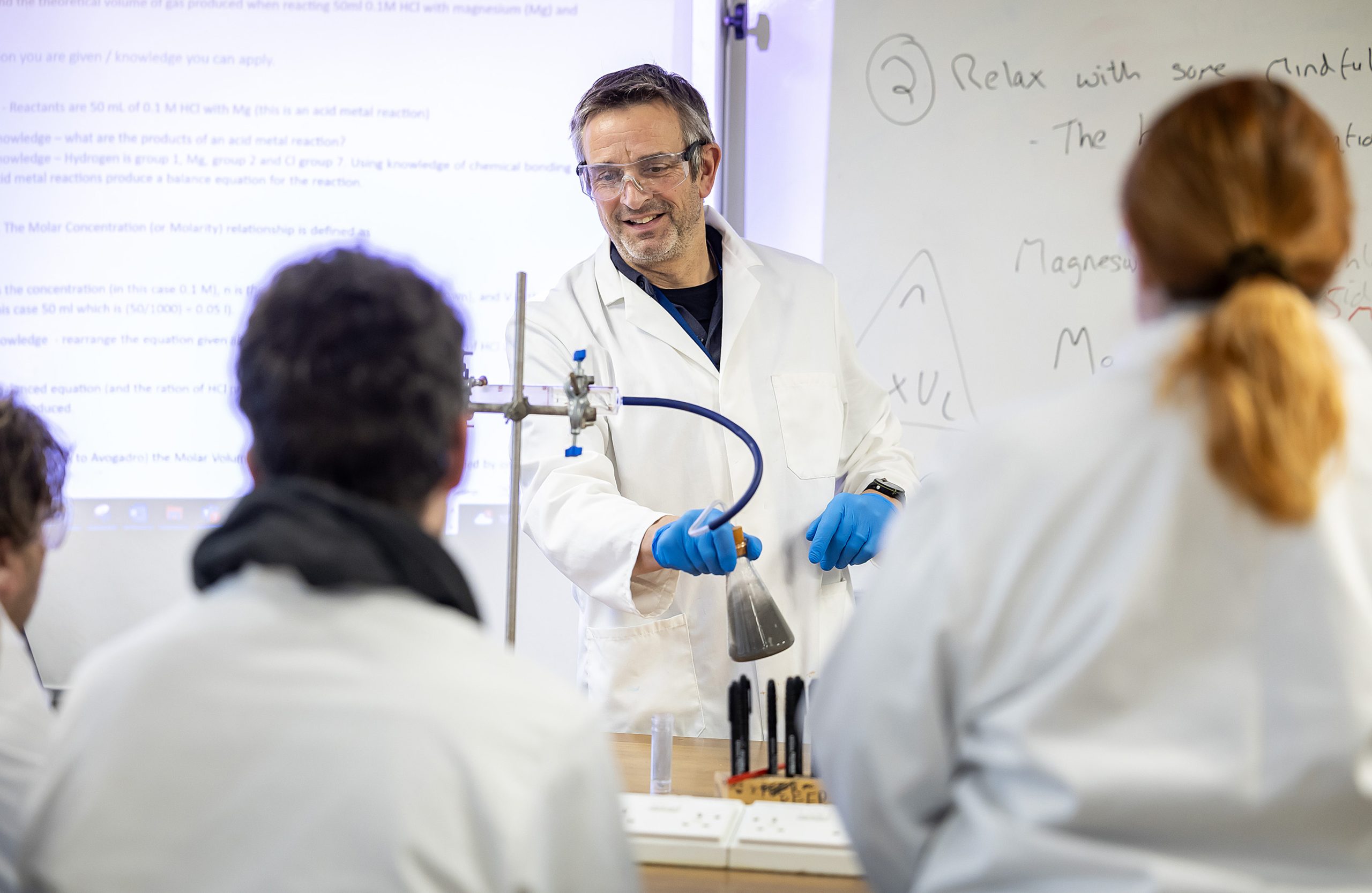 A lecturer undertakes a scientific demonstration in a lab wearing a white lab coat and holding a beaker.